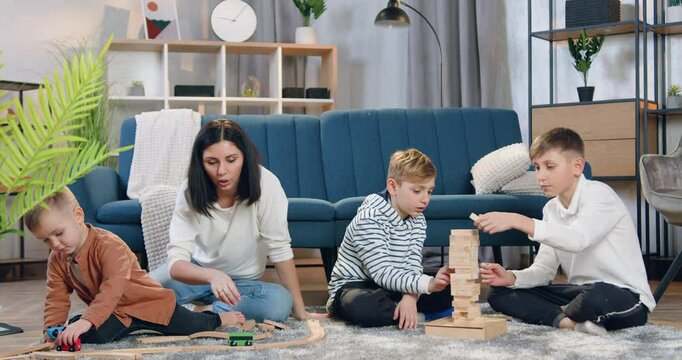 Boys playing with wooden blocks and demolish the built tower while their loving pretty mother playing with their smallest brother with car on car track