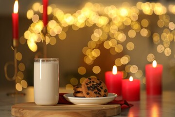 Glass of milk, tasty chocolate chip cookies and burning candles on table against blurred Christmas lights. Bokeh effect