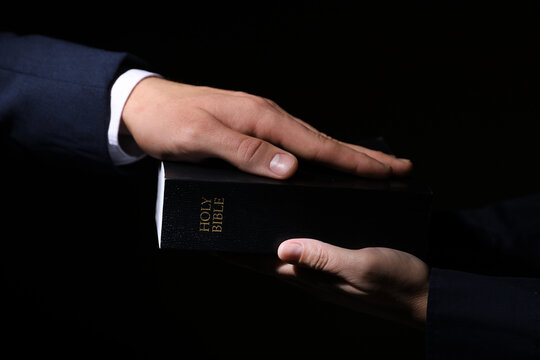 Man taking oath with his hand on Bible against black background, closeup