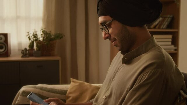 Side tilt shot of Indian man in black head hugger, glasses and kurta messaging on phone, sitting on couch at home