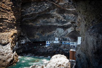 The image shows small white buildings with blue accents nestled within a large rocky cave by the coast. The vibrant blue-green ocean surrounds the rugged rocks, creating a picturesque scene.