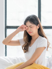 Young Asian woman crying and wiping her tears, expressing sadness and emotional distress while sitting on a bed near a window in a bright, quiet room