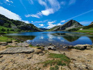 A tranquil mountain lake with clear, calm water, reflecting the blue sky and clouds. Surrounded by rocky hills and lush greenery. The foreground features a rocky and grassy shoreline.