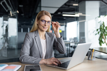 Confident businesswoman seated at a desk in a contemporary office environment, using a laptop. She adjusts her glasses while smiling, creating a friendly and professional atmosphere in the workspace.