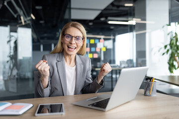 A joyful businesswoman in a gray suit celebrates a successful achievement at her office desk. Her...