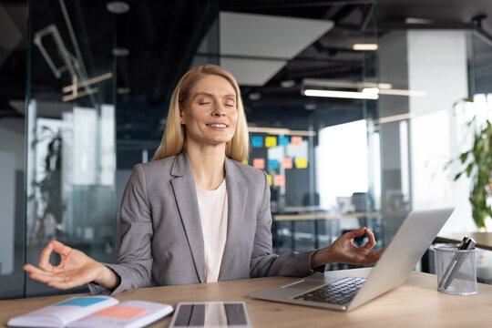 A businesswoman in a gray suit practicing mindfulness meditation at her office desk, promoting relaxation and stress relief during a busy workday. She is balancing work and well-being effectively.