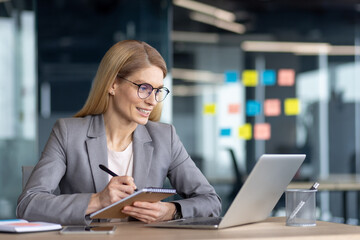 A professional woman in a suit is using a laptop in a modern office, jotting down notes with a smile, showcasing confidence and productivity during a meeting or work session.