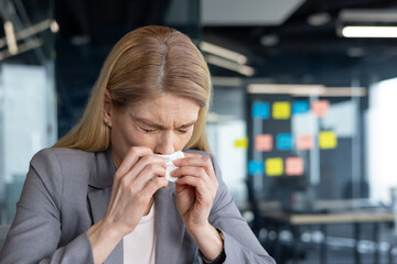 A professional woman in a business suit sneezes into a tissue at her office desk. The setting suggests a typical workday and highlights relatable health issues faced by working professionals.