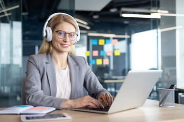 A professional woman wearing headphones works on her laptop at a desk in a modern office. She appears focused and engaged, exemplifying productivity and communication in a business environment.