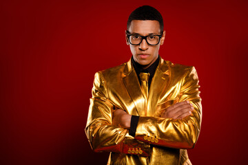 Young man in a striking golden suit crossing arms with a confident expression against a bold red...