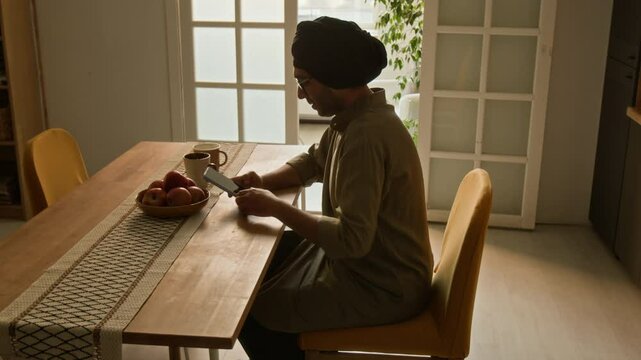Side shot of Indian man in traditional kurta and turban taking seat at dining table and having phone video call in domestic kitchen