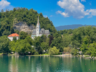 Fototapeta premium Lake Bled, Slovenia - June 28, 2024: St. Martin Church with gray-white mosaic roof surrounded by green foliage under blue cloudscape. White cliffs in back. Large white parish residential and office ho
