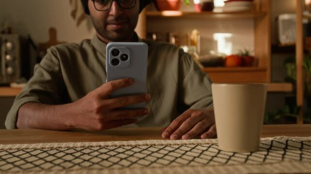 Tilt warm light shot of smiling Indian man in eyewear and traditional black turban sitting at dining table in domestic kitchen and texting messages on smartphone, communicating or surfing net