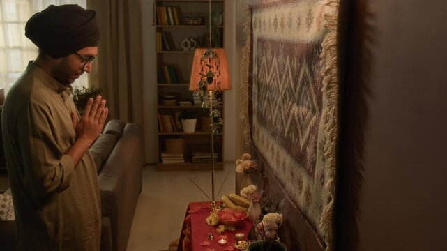 Side shot of young adult Indian man in turban and kurta worshiping by home Hindu altar, ringing bell and praying to statue of Ganesha, traditional ceremony