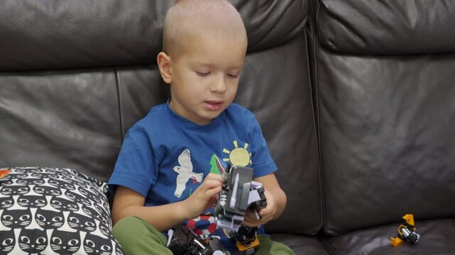 Child playing with new toy sitting on sofa at home. Concentrated face of little boy holding transformer robot toy. Three year old child examining new toy comments on his actions.