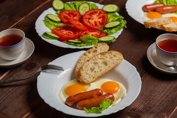 Breakfast two plates fried eggs with sausages white bread and tea in cups on a wooden table
