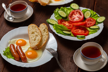 Breakfast fried eggs with sausages white bread and tea in a cup on a wooden table.