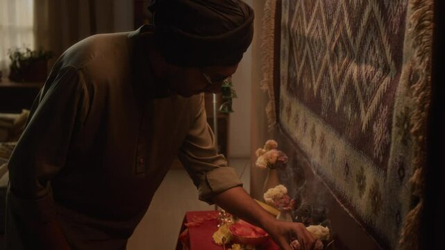 Side tilt shot of Indian man of 30s in turban and kurta praying by home Hindu altar, ringing bell and fixing smouldering incense sticks, traditional ritual, warm light