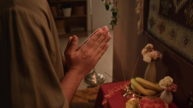Side tilt closeup of young adult Indian man worshiping to deities with namaste sign, reading prayers standing by home Hindu altar, traditional religious ceremony, warm light