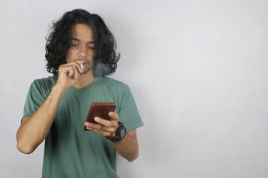 A young man smokes while looking down at his smartphone, appearing focused and distracted as smoke rises around his face.