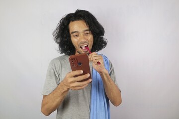 A man brushes his teeth while focusing on his smartphone, holding a towel on his shoulder. The image reflects a modern, multitasking morning habit.