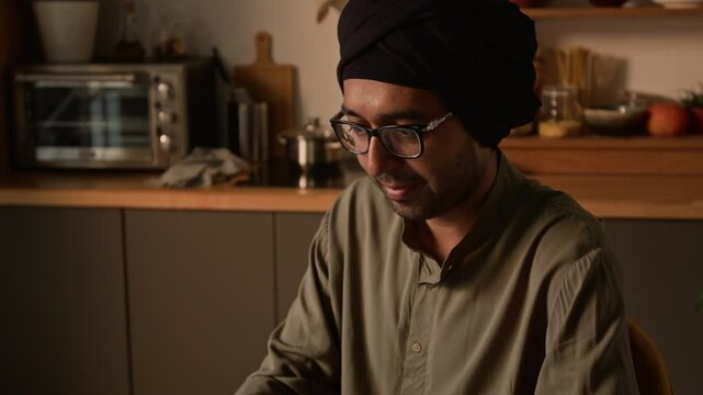 Tilt shot of male Indian programmer in glasses and turban coding on laptop sitting at dining table, working remotely from home