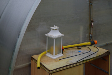 A white lantern on a wooden surface with a yellow garden hose and black cables, set inside a greenhouse with a metal frame