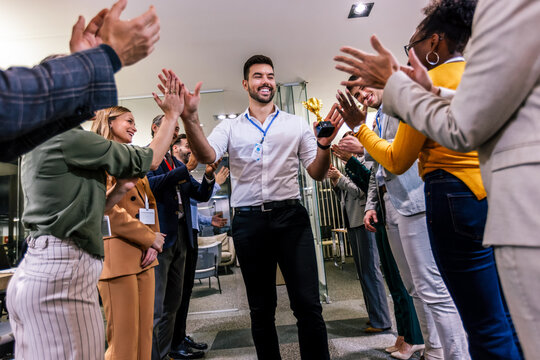 Young businessman receiving prize cup, proud colleagues applauding for him and cheering.