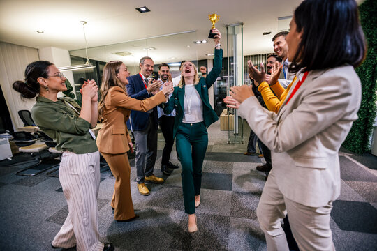 Young businesswoman receiving prize cup, proud colleagues applauding for her and cheering.