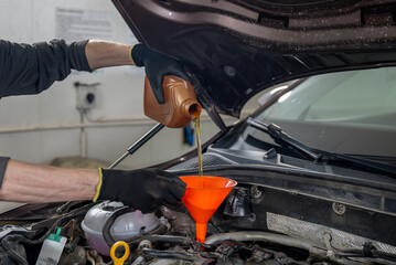 Fototapeta premium Mechanic in gloves pouring motor oil into a car engine using an orange funnel, in a workshop. Concept of vehicle maintenance 