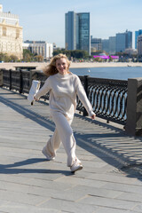 Full length side view portrait of beautiful playful long haired blond caucasian woman in white clothes walking on city street embankment in a sunny autumn day. Soft focus. Fashion and happy people.