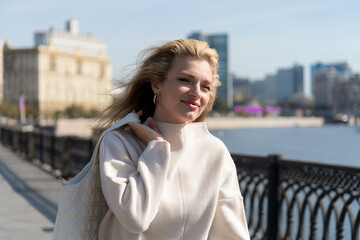 Portrait of beautiful smiling long haired blond caucasian woman in white clothes walking on city street embankment in a sunny autumn day. Clear blue sky. Soft focus. Fashion and happy people theme.