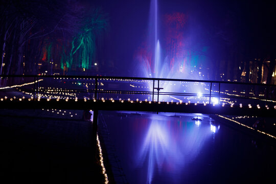 Nighttime canal scene, illuminated by colorful fountain and string lights.  Bridge spans the water.  Vibrant colors.