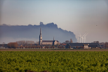 Large plume of smoke hangs over rural Dutch landscape, obscuring church and farm buildings.