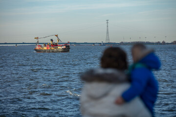 River scene with a decorated ship, bridge, and wind turbines.  People stand on the shore.