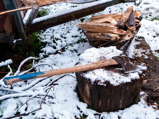 The axe lies on a wooden stump with a pile of chopped firewood on the snow-covered ground. Heating the house with wood.