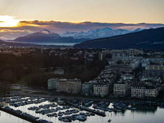 Breathtaking panoramic sunrise over Geneva city with Lake Geneva, moored boats, buildings, and snow capped Alps mountains in the background