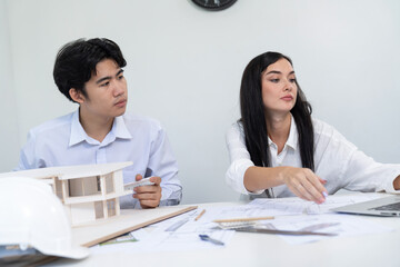 Professional male asian architect using ruler to measure house model length while young beautiful caucasian colleague using laptop to analyzed data on meeting table with house model. Immaculate.