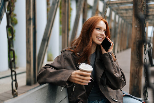 Stylish young redhead woman in leather jacket holding coffee cup and chatting on phone sitting on city swing at park on spring day. Happy red-haired female enjoy smartphone talking on park swing.