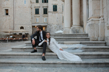 Portrait of young bride in white dress and groom in suit sitting on stairs near old city house in Croatia. Beautiful and romantic wedding, happy couple. enjoying the wedding day ceremony. Marriage.