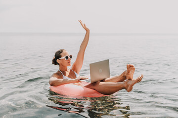 A woman is sitting on a pink inflatable raft in the ocean, holding a laptop. She is smiling and she is enjoying her time. Concept of relaxation and leisure.
