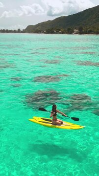 Vertical Screen Video. Aerial drone shot woman paddle boarding coral reef tropical island. Young female rows sitting on yellow paddleboard in crystal turquoise sea water. Outdoor lifestyle travel