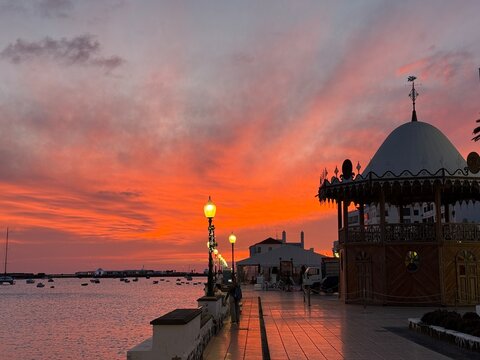 Atardecer pintoresco con colores vibrantes, paseo mar&iacute;timo, farolas encendidas y barcos anclados