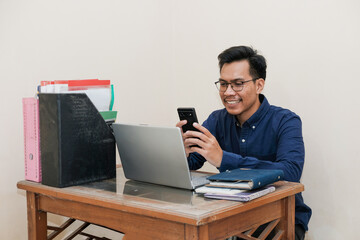 Smiling Southeast Asian Man Working While Sitting In Front Of Laptop In Home Office with hand holding handphone. Workplace Of Professional Worker, Freelancer or Student.