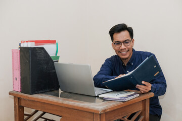 Smiling Southeast Asian Man Working While Sitting In Front Of Laptop and open notepad In Home Office. Workplace Of Professional Workers, Freelancers or Students.