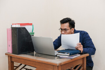Shocked Southeast Asian Man Works while Sitting in front of a laptop and holding paperworks at Home office. The Workplace of a Professional Worker, Freelancer or Student. Distance Work Concept.