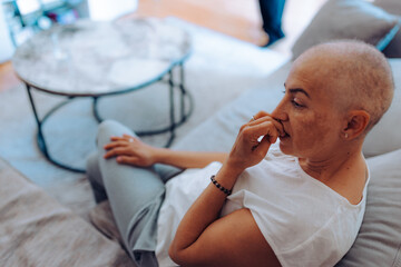 A thoughtful woman with cancer sits contemplatively on a couch biting nails, presenting an introspective moment filled with emotional depth and reflection in a modern living room context.