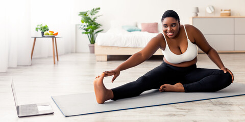 Plump black lady stretching legs, practicing yoga on mat, following tutorial on laptop, doing exercises online at home. Plus size African American woman trying to lose weight indoors