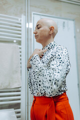 A bald woman thoughtfully adjusts her stylish outfit while preparing in a modern bathroom, showcasing her confidence and self-care routine amidst a bright setting.