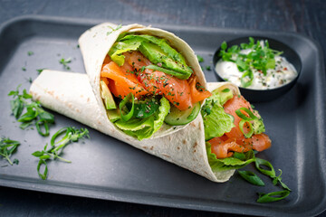Italian style wrap piada tortilla with cured graved salmon, salad and avocado served as close-up with sour cream dressing dip on a tray